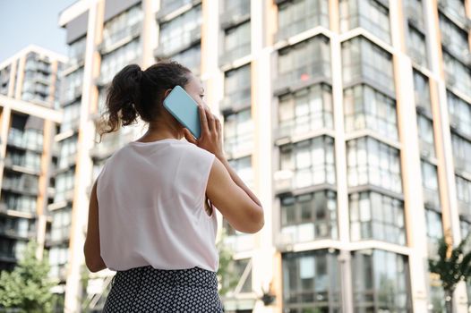 View from the back of a young African American, Hispanic mixed race woman talking on mobile phone on the background of urban high buildings. Business and communication concepts