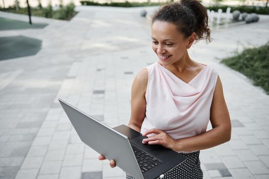 Gorgeous attractive Hispanic young businesswoman cutely smiling with toothy smile while working on laptop, standing on the urban buildings background. Business and communication concept