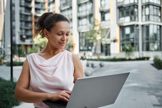 Gorgeous Hispanic young woman working on laptop, cutely smiling with toothy smile while looking at the screen monitor, standing on the urban buildings background. Business and communication concept