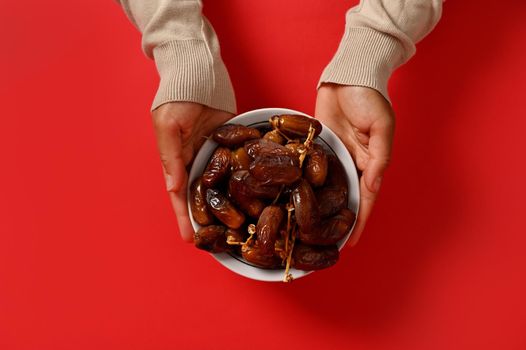 Flat lay composition of hands holding a delicious and sweet plate full of fresh dates, isolated on red background with copy space