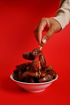 Female hands holding a branch of fresh tasty sweet dates. Vertical shot on red background with copy space. Dates for breakfast after interruption and fasting in Ramadan