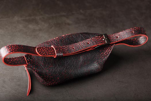 Belt bag made of textured, spotty black and red leather on a stone dark background. Elegant black and red bag with a zipper. Genuine leather, handmade. Banana bag