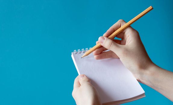 A hand with a pencil writes in a notebook on a blue background. Place for your text.