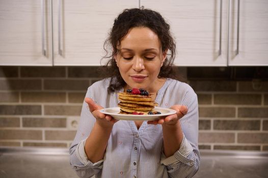 Charming woman holding a plate of delicious homemade freshly baked pancakes garnished with wild berries. Beautiful young dark-haired curly housewife with cooked pancakes for Shrovetide. Shrove Tuesday