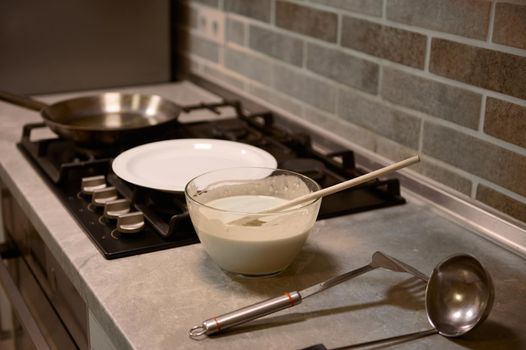 Wooden spoon in glass bowl with pancake batter, stainless steel kitchen utensils on kitchen countertop, white plate and frying pan on a black stove