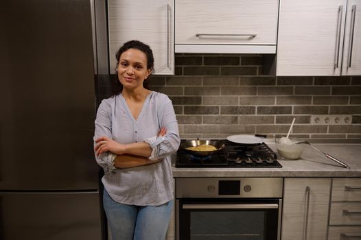 Charming young dark-haired curly pretty woman, housewife, smiling cutely at the camera, standing with her arms crossed at the stove and preparing homemade delicious pancakes for Shrovetide