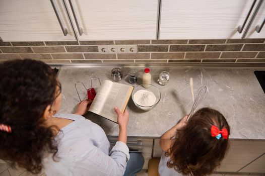 Overhead view of mother and daughter cooking food together while standing at the kitchen countertop. Mom holding recipe book ready to teach her adorable baby girl how to cook dough. Family time