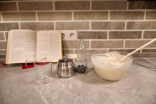 Transparent glass bowl with wooden spoon dropped into a liquid pancake dough, blueberries in a glass container on the background of a recipe-book on a kitchen countertop.