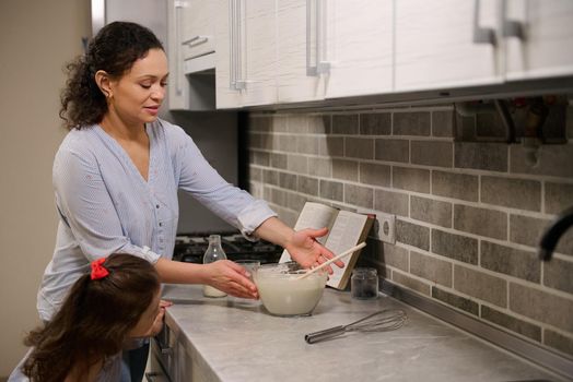 Mother and daughter enjoying spending time in the kitchen while cooking together. African American woman and cute little girl preparing liquid pancake dough for Shrove Tuesday.