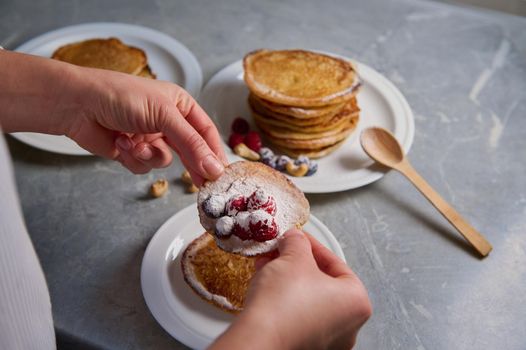Overhead view of the hands of female chef, housewife with homemade pancakes sprinkled with icing sugar on a white plate. Food background, cooking on Shrovetide, Shrove Tuesday concept