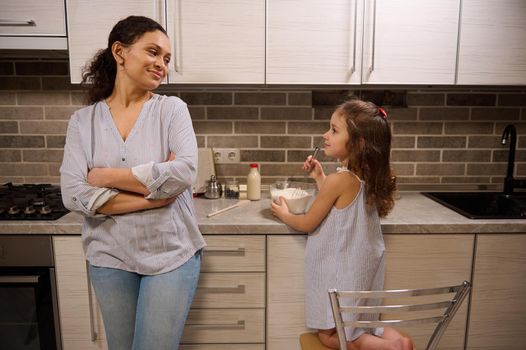 Beautiful smiling woman, happy loving mother leaning at kitchen countertop and admiring her adorable daughter cute baby girl mixing ingredients in a glass bowl while learning kneading pancakes dough