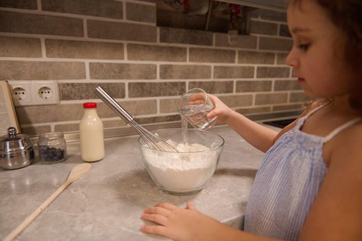 Soft focus on the hand of adorable little child girl, holding a glass with water and pouring it into a bowl with flour for preparing liquid dough for Shrove pancakes at home kitchen.