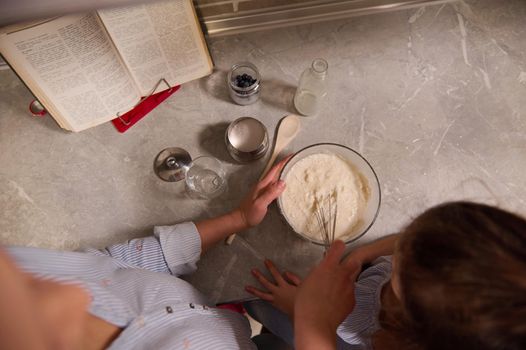 Overhead view of chef, woman, happy loving mother and her adorable daughter preparing pancake dough, mixing ingredients in bowl with whisk on kitchen countertop. Shrove Tuesday, food, culinary concept