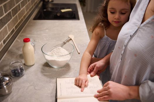 Beautiful happy child, adorable little girl points with her finger on a recipe book, standing at kitchen countertop close to her mom while enjoying cooking together, preparing dough for Shrove Tuesday