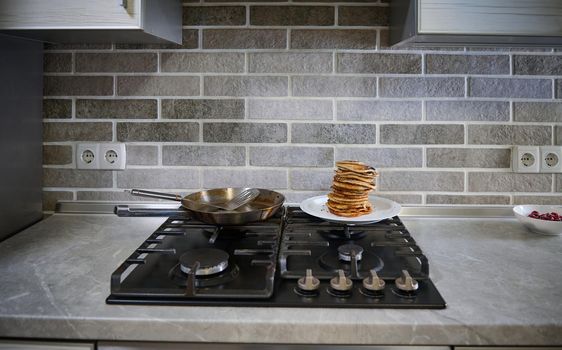 Front view of freshly baked homemade pancakes on white plate, steel frying pan and spatula on a black stove in the home kitchen. Shrove Tuesday concept