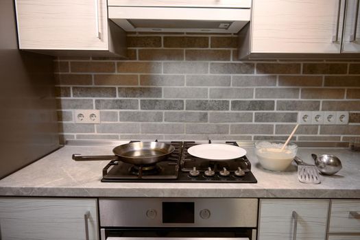 Wooden spoon in glass bowl with pancake batter, stainless steel kitchen utensils on kitchen countertop, white plate and frying pan on a black stove. Front view