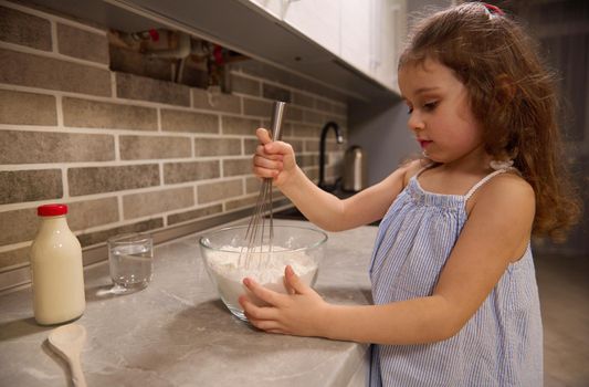 Adorable Caucasian little girl in blue dress mixing flour with dry ingredients ina glass bowl using a whisk, preparing pancake dough in the home kitchen for Shrove Tuesday