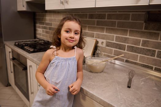 Portrait of a happy adorable beautiful little European girl in blue dress smiling cutely looking at camera, leaning her arm on a kitchen countertop while preparing dough at home kitchen