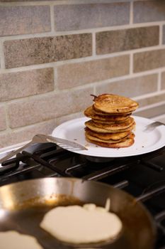 A stack of freshly baked homemade pancakes on a plate next to a black stove. In the blurred foreground is a frying pan with liquid dough pancakes. Food background. Shrove Tuesday concept