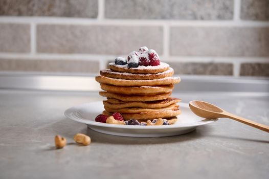 Stack of delicious homemade pancakes garnished with berries and cashew nuts, sprinkled with icing sugar on a white ceramic plate with wooden spoon on kitchen countertop. Culinary for Shrove Tuesday
