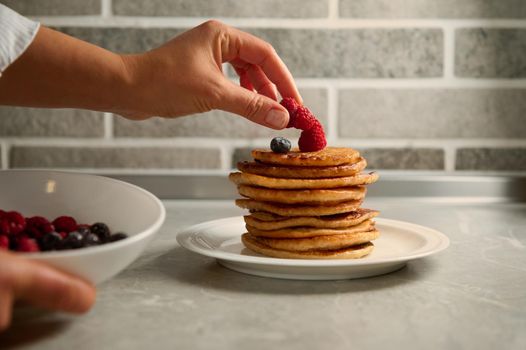 The chef's hand puts raspberries on a stack of delicious homemade freshly baked pancakes served on a white ceramic platter. Shrove Tuesday concept, food art with copy space for advertising
