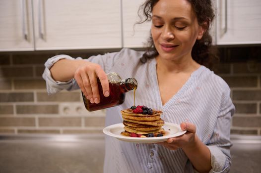 Focus on the hands of blurred beautiful female chef, charming pretty woman pouring maple syrup on a stack of homemade pancakes garnished with berries and cashew nuts. Shrove Tuesday concept, food art.