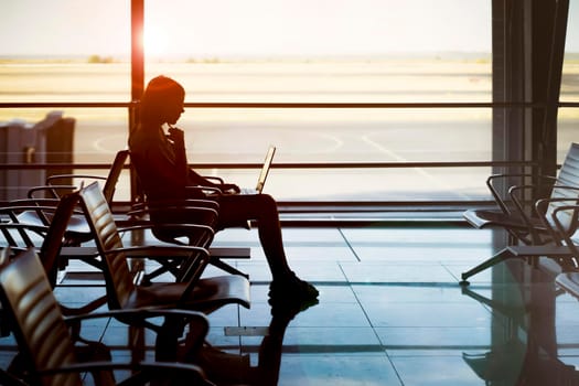 A young girl works on a laptop at the airport while she waits to board a plane. A woman buys tickets, studies and communicates via the Internet, the airport in the background.