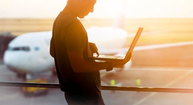 A young man is working on a laptop at the airport while waiting to board the plane. A man is engaged in business, buys tickets, studies and communicates via the Internet at sunset.