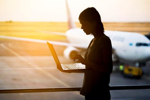 A young girl works on a laptop at the airport while she waits to board a plane. A woman buys tickets, studies and communicates via the Internet, the airport in the background.
