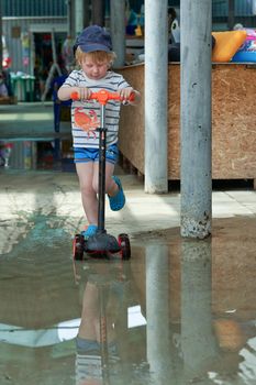 A child on a scooter rides through a puddle. Biking in a puddle