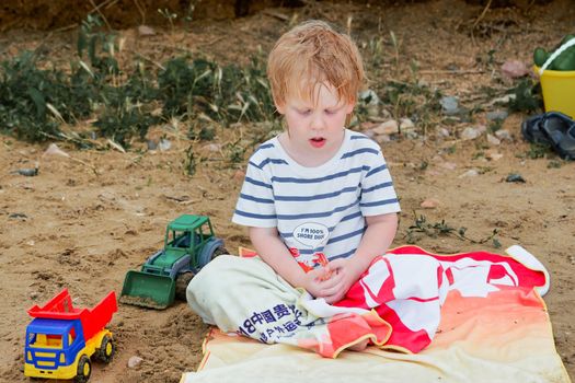 A little boy sits on a towel on a sandy beach among toys. Childrens holiday at the sea