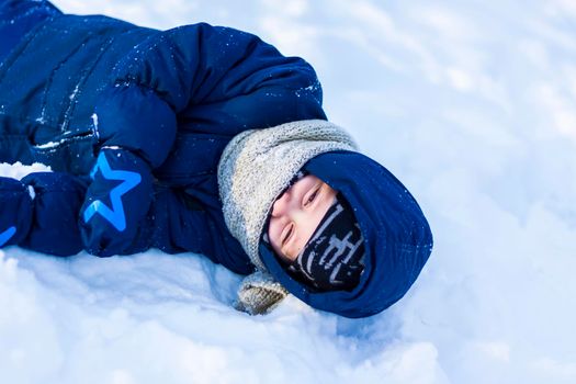 A smiling happy child lies in a snowdrift on a sunny winter day. A lot of snow and very frosty. Active winter outdoor games. The concept of a happy Christmas vacation.