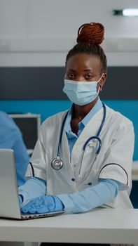 Portrait of young medic using laptop technology on desk in healthcare office at clinic. Doctor working with modern gadget in medical cabinet wearing white coat, gloves and face mask