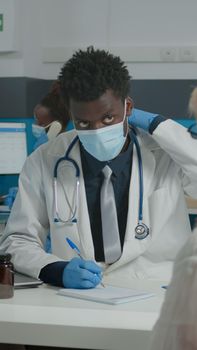 Medic with white coat sitting at desk while talking to elder woman in pain. Doctor doing healthcare checkup wearing face mask and gloves in medical cabinet with plexiglass wall at facility