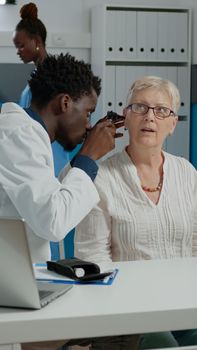 Otologist doing ear consultation for elder patient using professional otoscope at healthcare facility. Specialist with modern steel tool examining infection while sitting at desk in cabinet