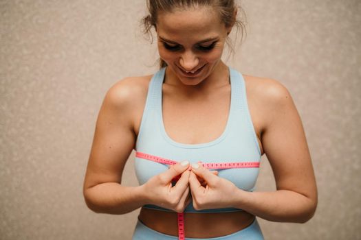 Cropped view of slim woman measuring breasts with tape measure at home, close up. A European woman checks the result of a weight loss diet or liposuction indoors. Healthy lifestyle