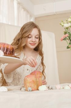 A girl with long hair in a light dress is sitting at the Easter table with cakes, spring flowers and quail eggs. Happy Easter celebration