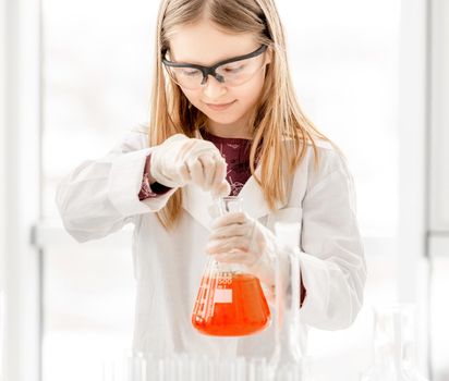 Girl doing scientific chemistry experiment wearing protection glasses and holding tube with orange water. Schoolgirl with equipment and chemical liquids on school lesson
