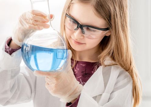 Smart girl doing scientific chemistry experiment wearing protection glasses, holding bottle and measuring blue liquid. Schoolgirl with chemical equipment on school lesson