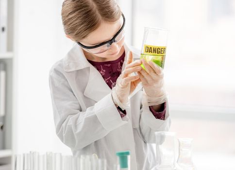 Smart girl doing scientific chemistry experiment wearing protection glasses, holding bottle with title danger. Schoolgirl with chemical equipment on school lesson