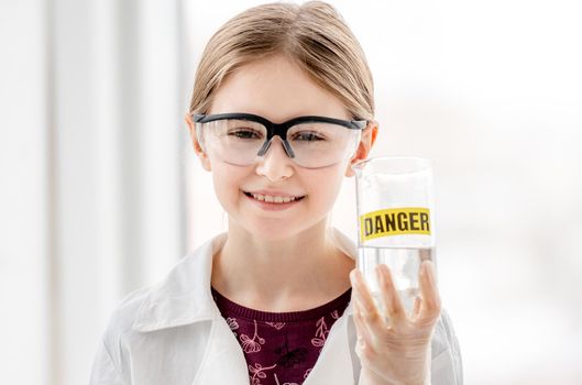 Smart girl doing scientific chemistry experiment wearing protection glasses, holding bottle with title danger. Schoolgirl with chemical equipment on school lesson