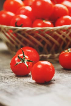 Fresh red tomatoes in a wicker basket on an old wooden table. Ripe and juicy cherry tomatoes with drops of moisture, gray wooden table, around a cloth of burlap. In a rustic style. Close-up vertical frame. Low contrast
