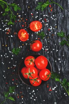Juicy red cherry tomatoes on a black background with spices, coarse salt and greens. Sweet and ripe tomatoes for salads and as ingredients for cooking. Top view, low contrast