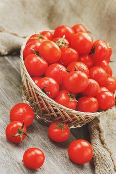 Fresh red tomatoes in a wicker basket on an old wooden table. Ripe and juicy cherry tomatoes with drops of moisture, gray wooden table, around a cloth of burlap. In a rustic style. Close-up vertical frame. Low contrast