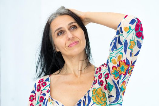 Close-up portrait of middle aged woman starting getting grey-haired wearing dress with flowers on white background, middle age sexy lady, cosmetology concept.