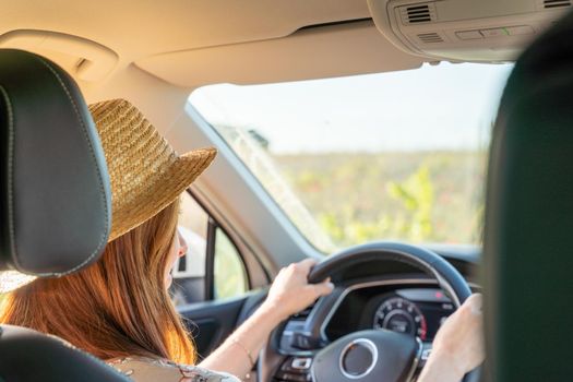 A woman in a hat and a colored dress is sitting at the wheel of a car, the view from behind, the window shows the field