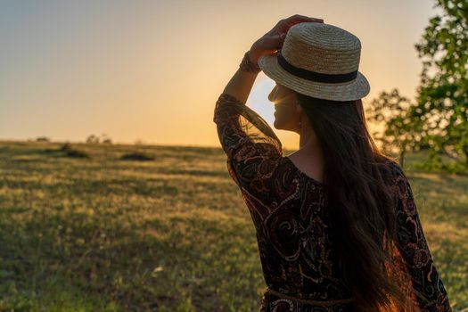 A happy woman travels alone in the forest at sunset, in a long dress, hat and a bouquet of flowers. Happy vacation. Rear view of a happy woman