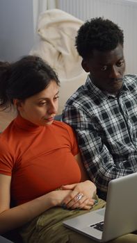 Young interracial couple talking on video call conference sitting on couch at home. Mixed race husband and wife using laptop for online remote communication in living room together