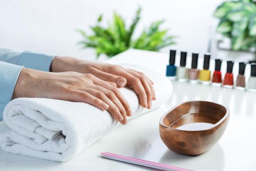 Female hands lying on white towel. Manicurist workspace with colorful nail polish bottles and wooden bowl with water. Female hand preparing for manicure. Professional nail care and beautician service.
