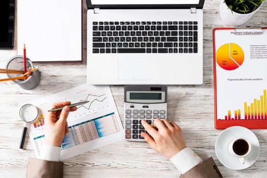 Businessman working at office desk with financial data. Top view office workplace with human hands using calculator. Finance accounting concept. Flat lay table with laptop and business charts.